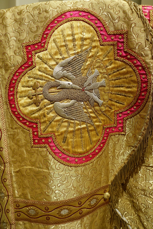 Nahaufnahme eines Priestergewandes mit aufwendiger Gold- und Rotstickerei und einem luxuriösen, königlich anmutenden Design vor einem neutralen Hintergrund.