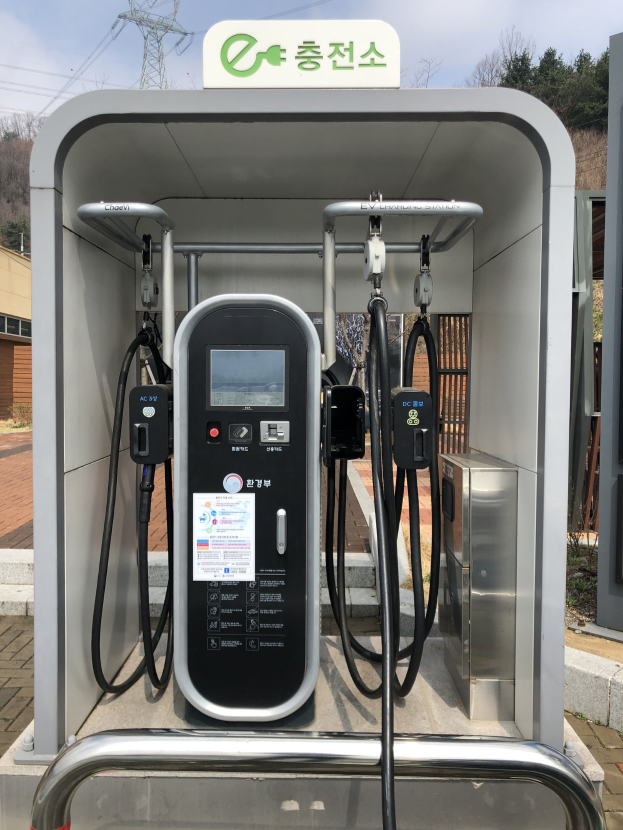 Elektrofahrzeug-Ladestation in Seoul, Südkorea, mit Ladegerät, verbundenen Kabeln und Schild, vor Gebäuden, Bäumen und Strommasten