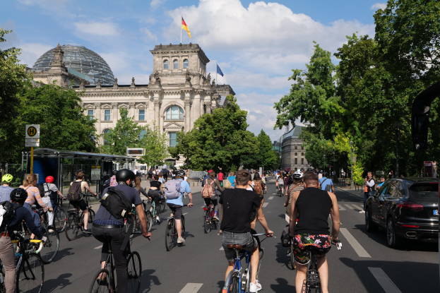 Gruppe von Menschen, die auf Fahrrädern eine von Bäumen gesäumte Straße in Berlin, Deutschland, entlangfahren, mit Gebäuden und einer Bushaltestelle im Hintergrund, unter einem bewölkten Himmel und einer Flagge, die auf einem Gebäude weht.