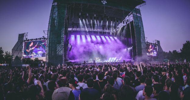 Eine große Menge an einem Musikfestival schaut auf einer hell beleuchteten Bühne mit Instrumenten und Bildschirmen, die vor einem Gebäude, Bäumen und einem klaren blauen Himmel liegen.