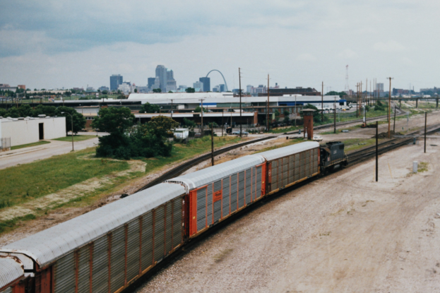 Güterzug, der auf Schienen neben einer Stadtlandschaft mit Strommasten, Kabeln, Gebäuden, Bäumen, Gras und einem bewölkten Himmel fährt.