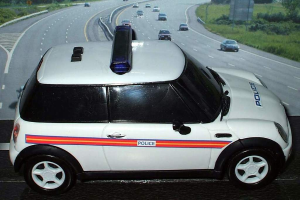 Ein weißer Polizeiwagen steht im Vordergrund mit mehreren Fahrzeugen, die sich auf der Straße dahinter bewegen, umgeben von Gras und Bäumen auf beiden Seiten in einer ländlichen Umgebung.