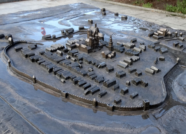 Eine Modellstadt in einer Pfütze umgeben von Gebäuden, Grünflächen und einer Wand im Hintergrund.