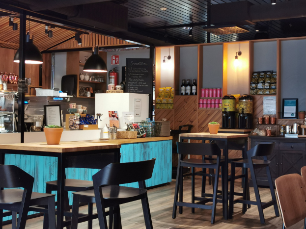 Innenansicht eines Restaurants mit Holztischen, Stühlen, einem Blumentopf mit einer Pflanze auf einem Tisch, einer Texttafel, einem Kühlschrank, einem Feuerlöscher, Regalen mit Flaschen, Schränken mit Gegenständen, einem gerahmten Foto an der Wand, Deckenleuchten und Lampen.