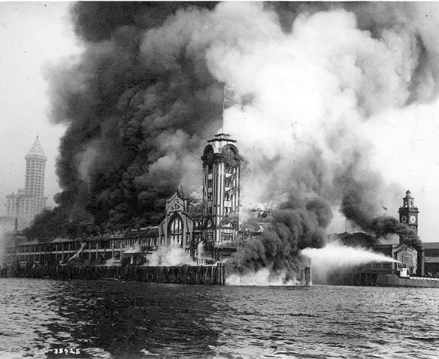 Schwarz-weiß-Foto eines großen Gebäudes, das in Flammen steht und von dem Rauch aufsteigt, mit einem Gewässer im Vordergrund und anderen Gebäuden und Himmel im Hintergrund.