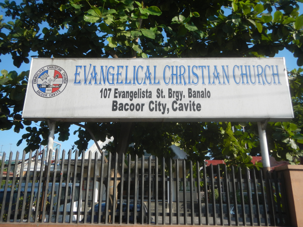 Eine evangelikale christliche Kirche in Bacoor City, Cavite, mit einem Schild, einem Metallzaun, einer Wand, Bäumen, Fahrzeugen, Gebäuden, Pfählen und einem bewölkten Himmel.