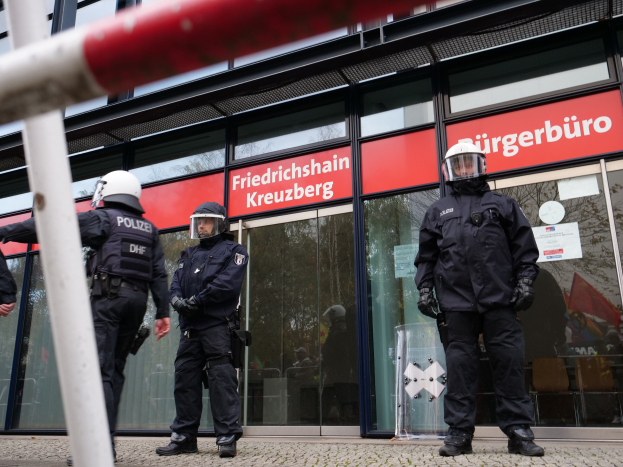 Polizeibeamte in Uniform vor einem Glaswand-Gebäude mit einem Pfahl links und Schildern mit "Friedrichshain Kreuzberg"-Text im Hintergrund.