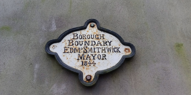 Schwarze Metalltafel an einem Gebäude mit weißer fetter Schrift, die "Borough Boundary Edm Smithwick Mayor" liest.