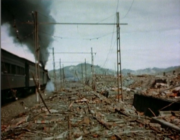 Ein Zug fährt durch ein beschädigten Gebiet mit Rauch, der aus ihm aufsteigt, umgeben von Strommästen und -dröhten, mit Höhen und einem klaren Himmel im Hintergrund und verstreuten Trömmern am Boden.