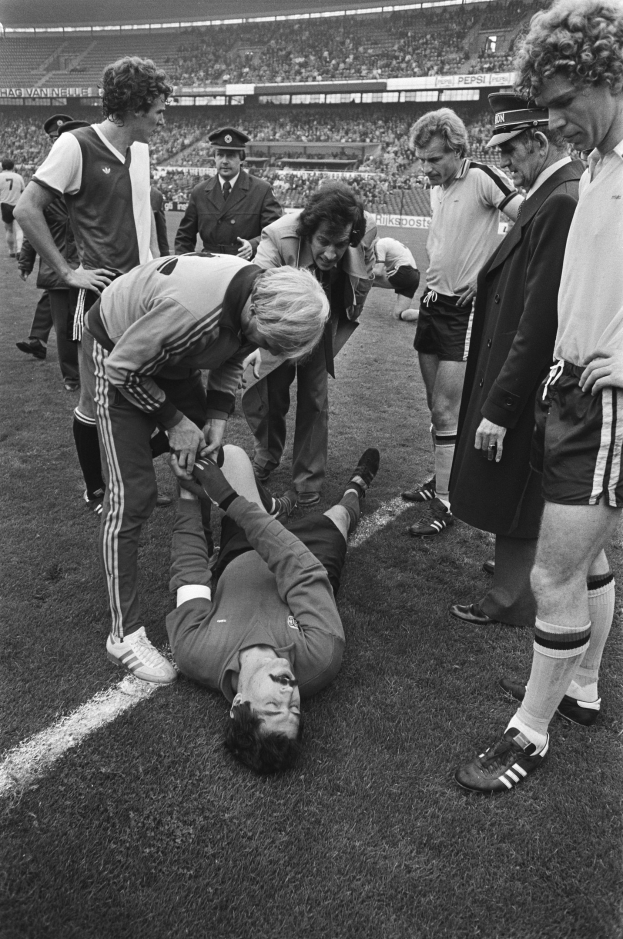 Schwarzes und wei├čes Bild eines fünfk├Ąmpfigen Spielers auf dem Boden umgeben von einer Gruppe von M├Ąnnern, mit einer Menge im Stadionhintergrund; der Spieler scheint verletzt zu sein.
