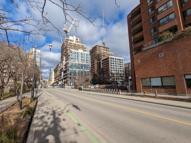 Eine Stadtstraße mit hohen Gebäuden, Straßenlaternen, Strommasten, Lampen, Kabeln, Fahrzeugen, Fußgängern, Geländern, Bäumen und einem bewölkten Himmel.