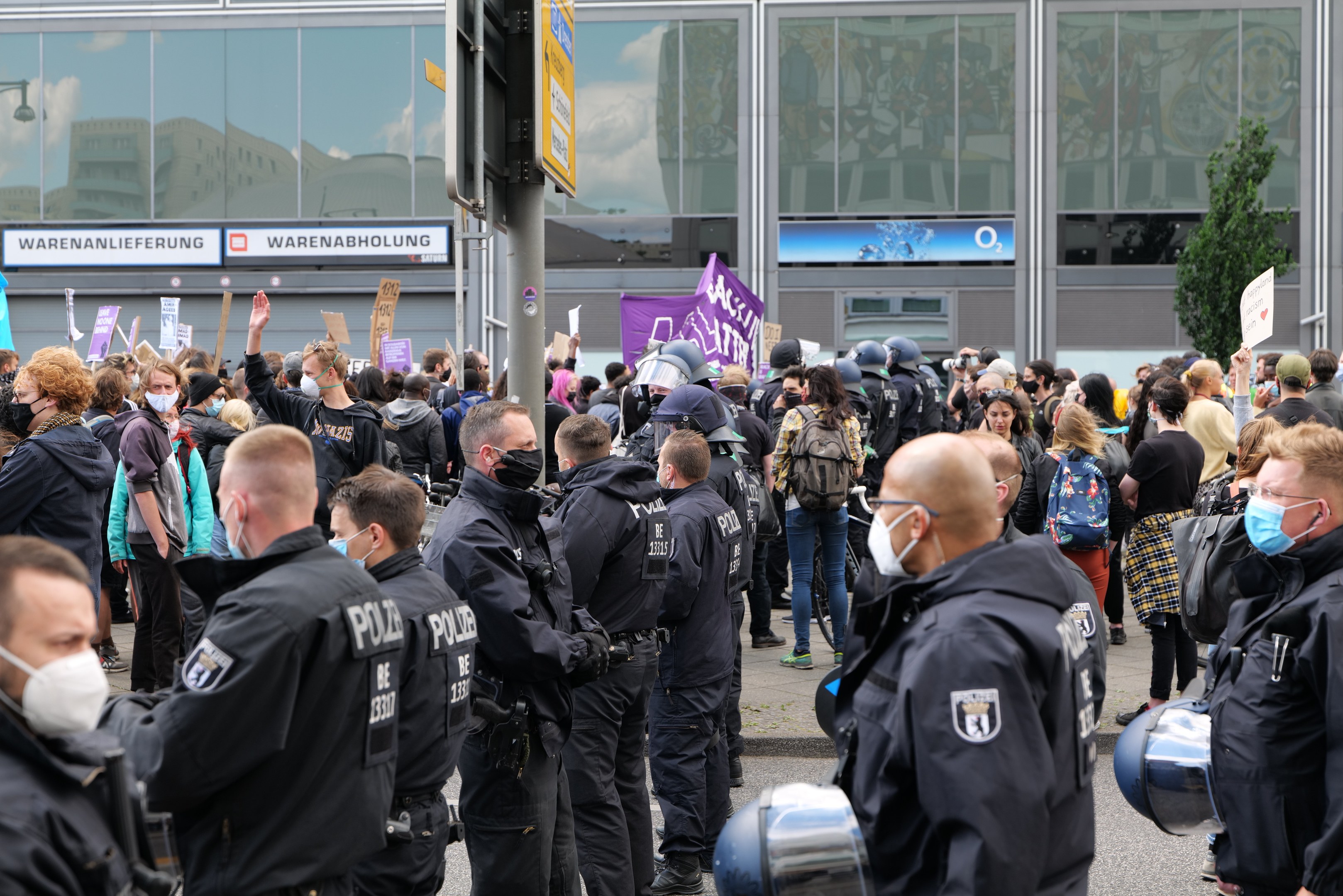 Eine große Gruppe von Menschen steht vor einem Gebäude, einige halten Schilder und tragen Helme, mit einem Schildständer im Vordergrund und einem Baum im Hintergrund, was auf eine Demonstration hindeutet.