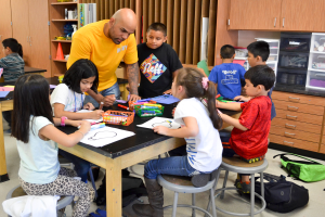 Eine Gruppe von Kindern sitzt um einen Tisch in einem Klassenzimmer, während ein Mann vor ihnen steht, umgeben von Papieren, Stiften und Taschen. Im Hintergrund sind Regale und Gestelle mit verschiedenen Gegenständen zu sehen.