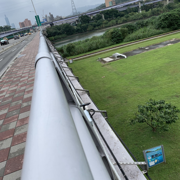 Stadtansicht von einem Gebäude mit einem großen Metallwasserrohr im Vordergrund, Straßen mit Fahrzeugen, Bäumen, Gebäuden und Versorgungsinfrastruktur gegen den Himmel.