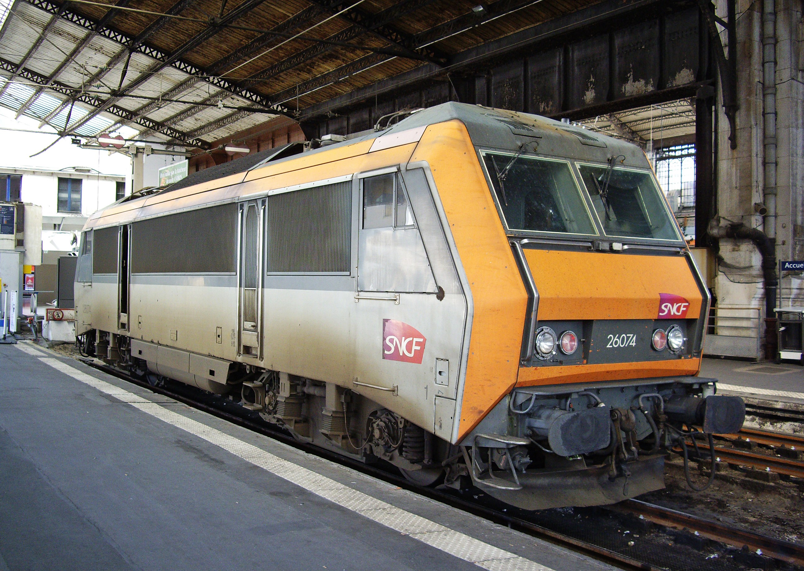 Ein dieselhydraulischer Lokomotive auf einem Bahnhof mit einem Bahnsteig auf der linken Seite und Gebäuden, Pfählen und anderen Objekten im Hintergrund.