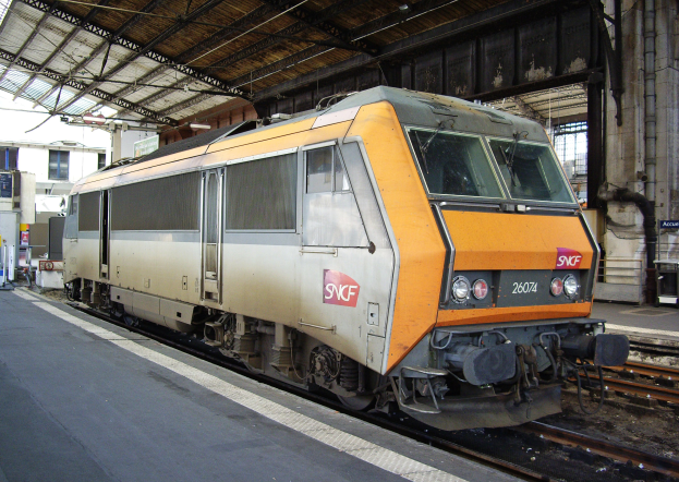 Ein dieselhydraulischer Lokomotive auf einem Bahnhof mit einem Bahnsteig auf der linken Seite und Gebäuden, Pfählen und anderen Objekten im Hintergrund.