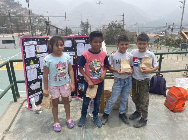 Gruppe von Kindern auf einem Basketballfeld mit Papiertüten in den Händen, Tüten und Gegenstände rechts und eine Tafel mit Text links, vor einem Hintergrund aus Zäunen, Pfosten, Straßenlaternen, Bäumen, Gebäuden, Hügeln und Himmel.
