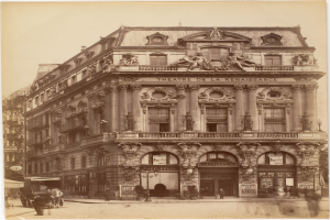 Ein altes Foto des Théâtre de la Renaissance in Paris, Frankreich, das ein Gebäude mit Fenstern, Säulen, Bögen und Skulpturen an der Fassade zeigt, mit ein paar Menschen und Fahrzeugen auf der Straße davor und dem Himmel im Hintergrund.