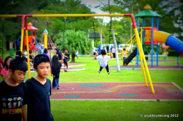 Kinder auf Spielgeräten im Park mit Bäumen im Hintergrund.