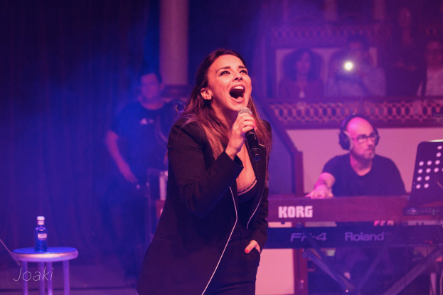 Frau singt in ein Mikrofon auf der Bühne, rechts ein Pianist und im Hintergrund Publikum, links auf einem Hocker eine Flasche.