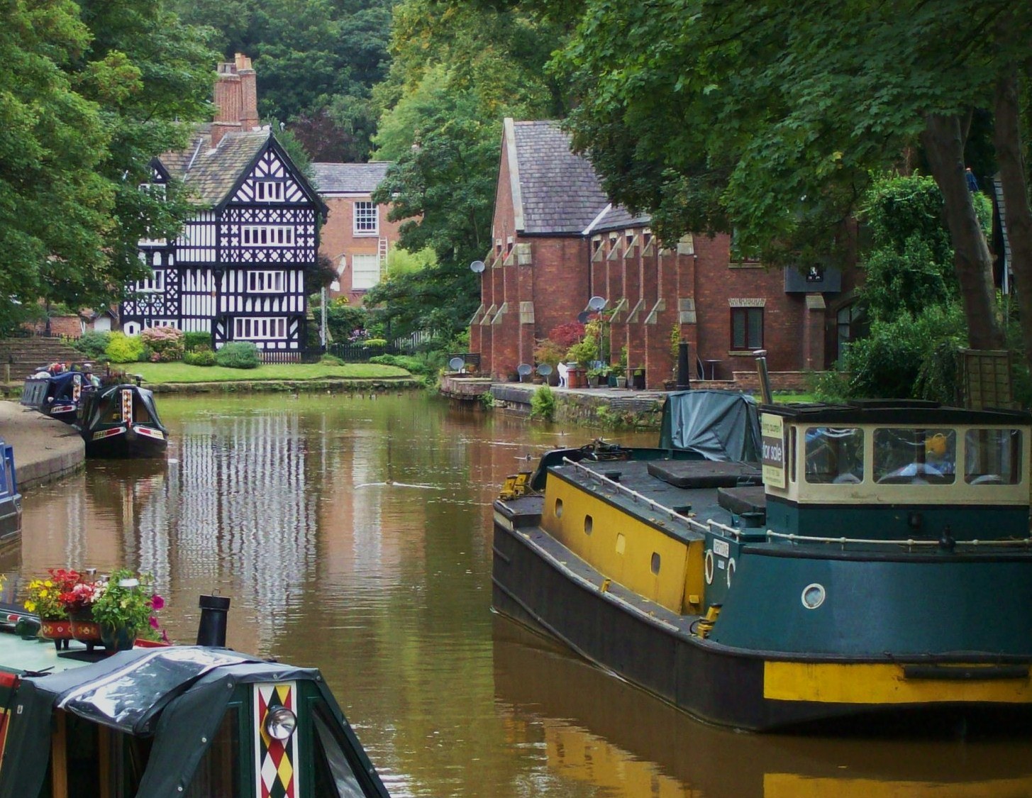 Kanal mit mehreren Booten, die an seinem Rand liegen, umgeben von grünem Gras, Pflanzen und Bäumen, mit Gebäuden mit Fenstern im Hintergrund.