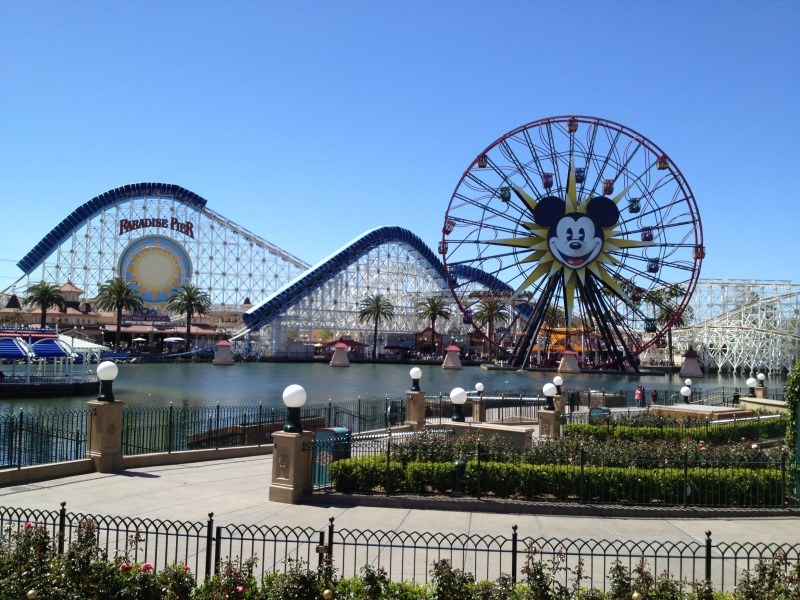 Der Vergnügungspark California Adventure im Disneyland mit einem Riesenrad, einer Achterbahn, Grünflächen, Laternenmasten, einem Gewässer und einem hellblauen Himmel.