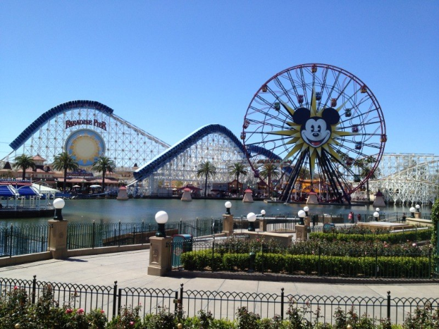 Der Vergnügungspark California Adventure im Disneyland mit einem Riesenrad, einer Achterbahn, Grünflächen, Laternenmasten, einem Gewässer und einem hellblauen Himmel.