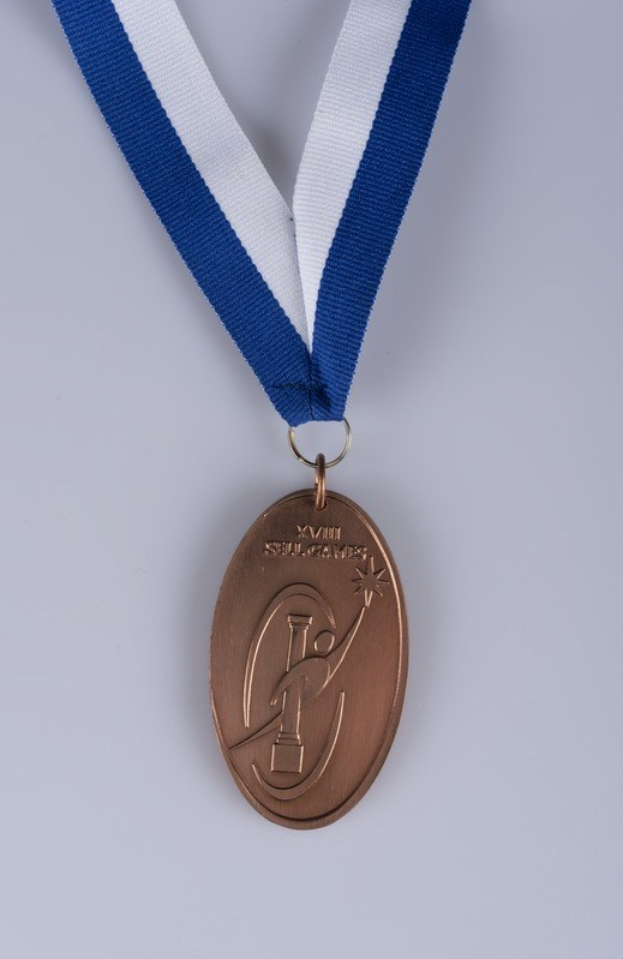 Goldene Medaille mit blau-weißem Band auf weißem Hintergrund, mit eingravierter Schrift.