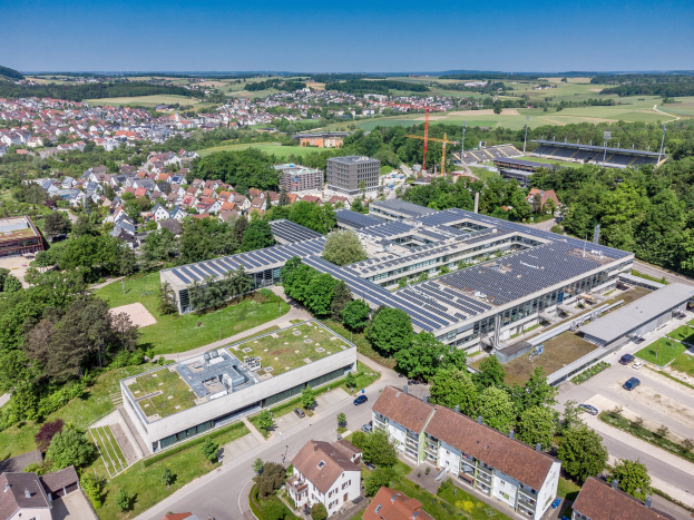 Luftbild einer großen Gebäudes mit Solarmodulen auf dem Dach, umgeben von Häusern, Bäumen, Fahrzeugen auf einer Straße, Hügeln im Hintergrund und einem klaren blauen Himmel.
