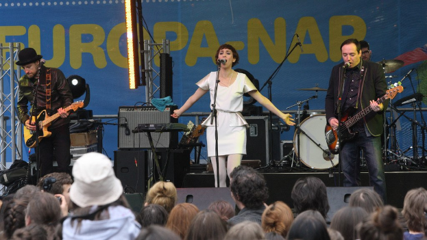 Vier Musiker auf der Bühne: Eine Frau in einem weißen Kleid singt in der Mitte, flankiert von zwei Männern mit Gitarren, mit einem Publikum darunter und Lautsprechern gegen eine Wand im Hintergrund.