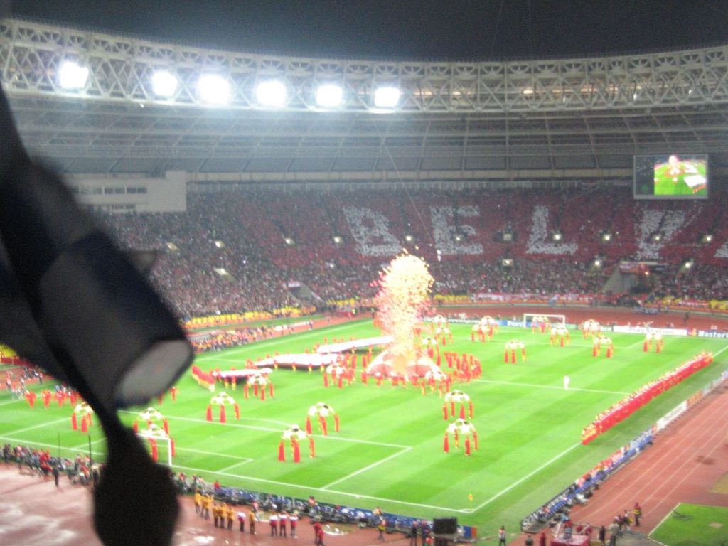 Ein Stadion voller Zuschauer, mit Flutlicht, Eisengittern, Werbetafeln, Bildschirmen, Überwachungskameras und verschiedenen Dekorationen einschließlich Fahnen.
