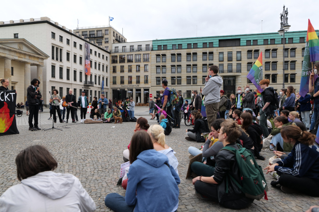 Eine Gruppe von Menschen, die auf dem Boden vor einer Menge sitzen, die Fahnen und Schilder hält, während einer Anti-Schwulen-Demo in Berlin. Im Hintergrund sind eine Statue, ein Mikrofonständer und Gebäude zu sehen.