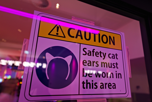 Hellgelbes Warnschild in einem Ladenschaufenster mit der Aufschrift "Achtung: Sicherheitskatzenohren müssen in diesem Bereich getragen werden." vor einem unscharfen Hintergrund.