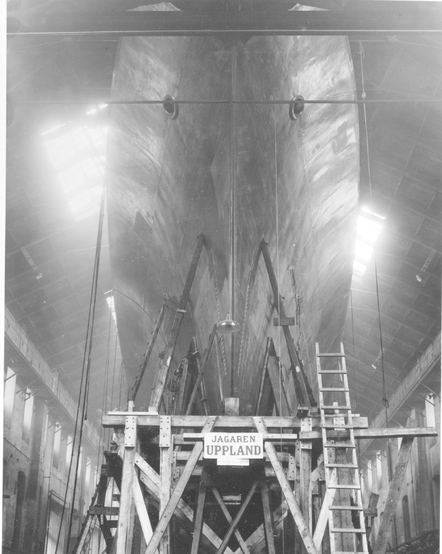 Schwarzes und weißes Foto eines großen Bootes im Bau in einer Werft, das eine Leiter, Holzpfosten, ein Schild, Metallrahmen, Deckenlampen und Fenster zeigt.