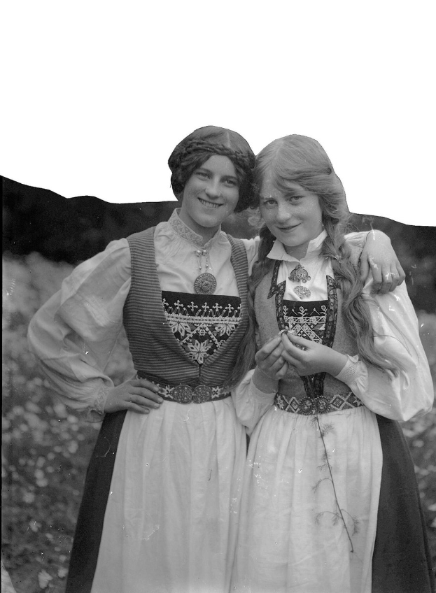 Zwei junge Frauen in traditionellen bayrischen Kleidern mit weißer und schwarzer Stickerei und Perlen stehen lachend vor üppigen grünen Pflanzen unter einem hellen weißen Himmel.