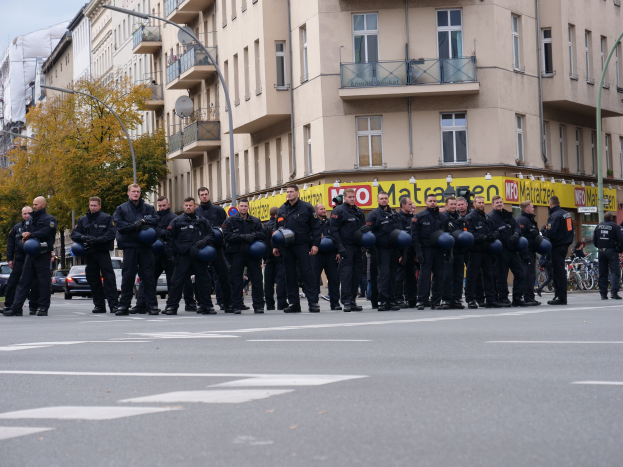 Eine Gruppe von Polizisten in schwarzen Uniformen mit blauen Helmen steht in einer Straße, die von Laternenmasten, Bäumen und Gebäuden gesäumt ist, unter einem klaren blauen Himmel.