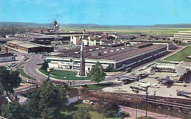 Altes Schwarz-Weiß-Foto eines Flughafens mit geparkten Flugzeugen, Gebäuden, Bäumen, Straßen, Fahrzeugen, Strommasten, Gras, Bahnschienen, Türmen, Hügeln und einem klaren blauen Himmel, mit Text entlang der unteren Kante.