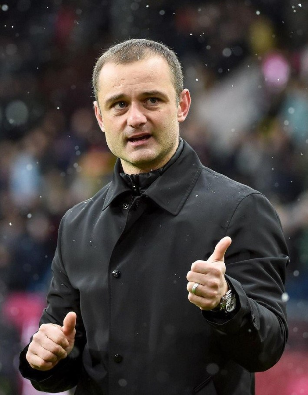 Ein Mann in einer schwarzen Jacke zeigt den Daumen hoch, tr├Ągt eine Uhr und hat einen unscharfen Hintergrund.