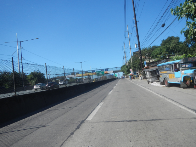 Straßenverkehr mit fahrenden Fahrzeugen, geparktem Bus, Strommästen mit Drähten, einem Zaun auf der linken Seite, Bäumen, Laternenmästen, einer Brücke im Hintergrund und einem blauen Himmel.