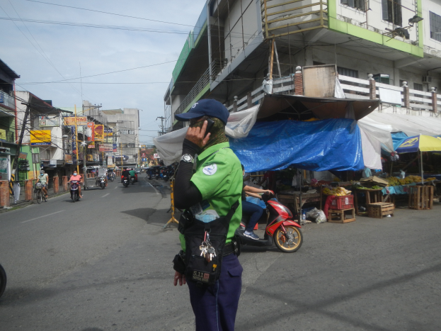 Ein Polizeibeamter in grüner und blauer Uniform und einer blauen Mütze steht auf der Seite einer Straße, spricht auf einem Handy, mit Fahrzeugen im Hintergrund, Ständen mit Gegenständen zu seiner Rechten und einem bewölkten Himmel darüber.