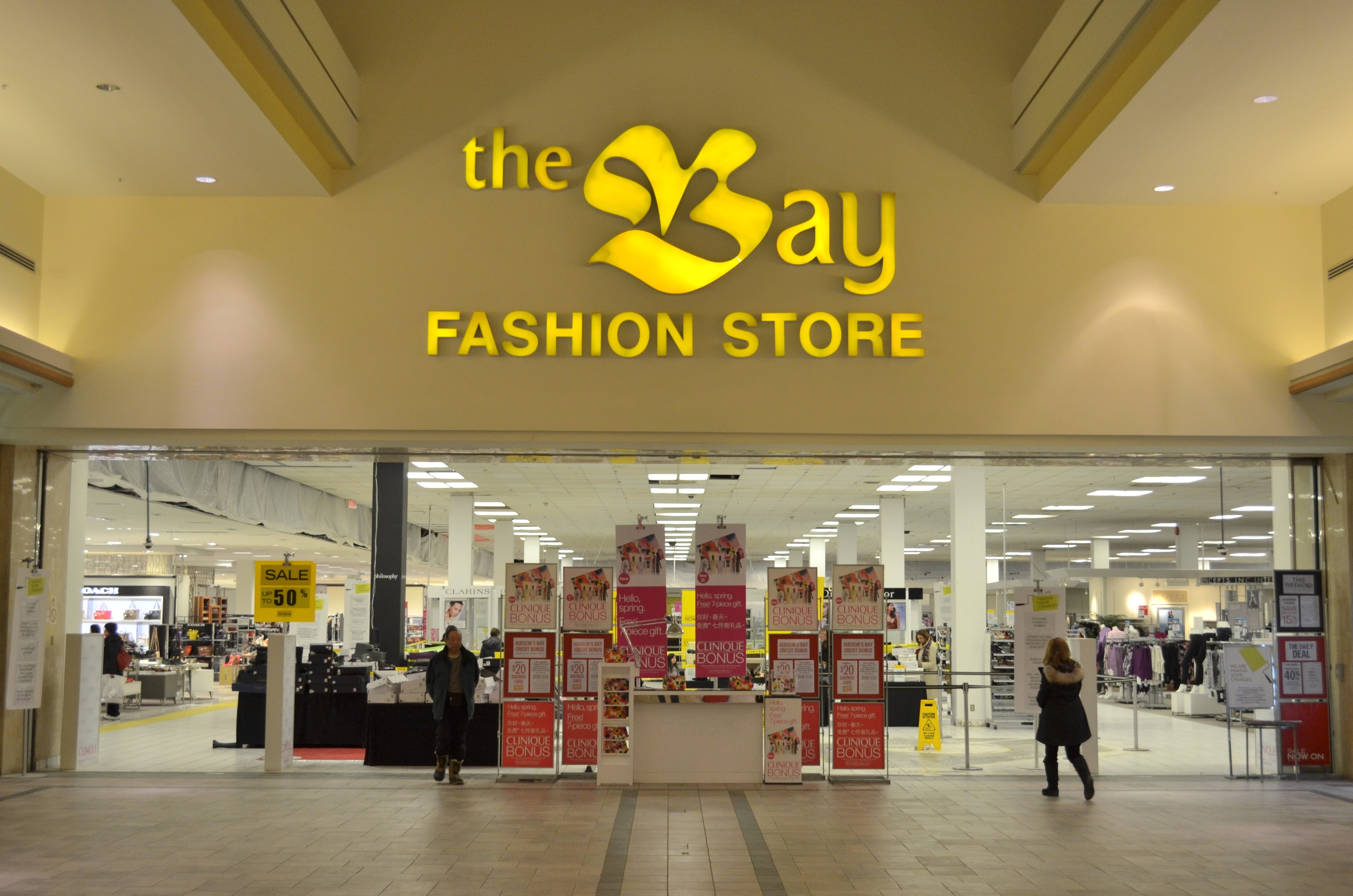 Verkaufsfläche eines Bay Fashion Shops in einem Einkaufszentrum mit Einkäufern, Wandtexttafeln, Säulen, Deckenleuchten und Hintergrundauslagen.