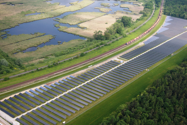 Luftaufnahme einer Solar-Farm mit Panels in einem grasbewachsenen Feld, umgeben von Bäumen und Wasser, mit einem Zug auf einer nahen Strecke.