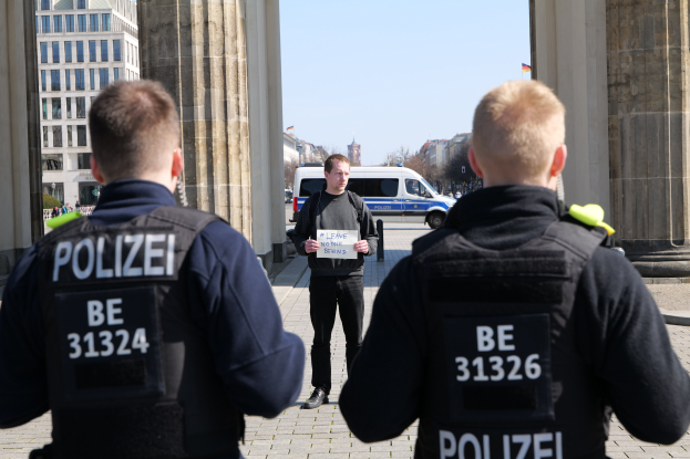 Drei Polizeibeamte stehen vor einem Gebäude, mit einem Mann, der ein Papier zwischen ihnen hält, flankiert von Säulen und vor dem Hintergrund von Fahrzeugen, Menschen, Bäumen, Gebäuden und Himmel.