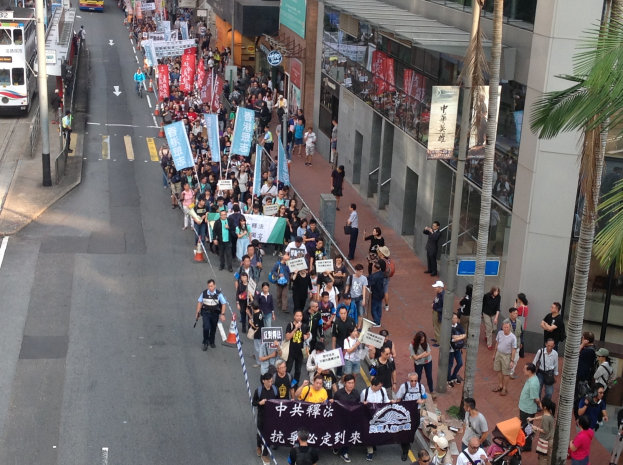 Eine große Gruppe von Menschen marschiert mit Protestschildern die Straße entlang, mit Bäumen, Glasfassaden, Fahrzeugen und Straßenlaternen und Schildern an der Seite.