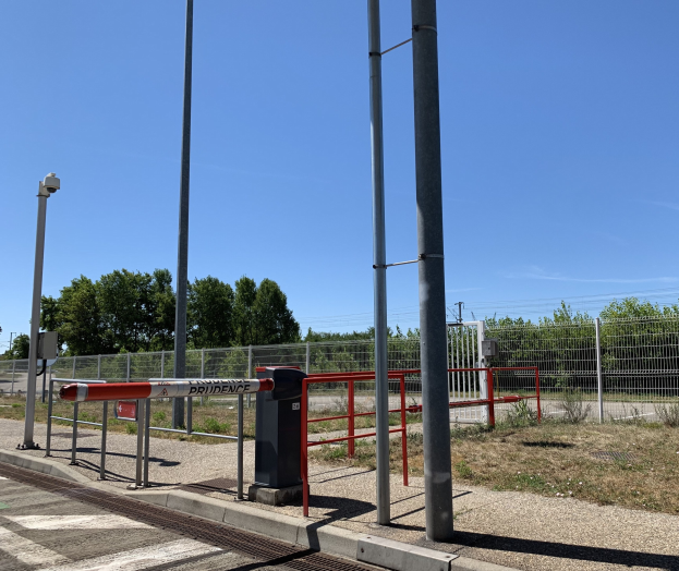 Ein rotes und weißes Schild an einem Bahnhof, umgeben von einem Metallzaun, Bäumen, Pflanzen, Gras, einem Straßenmast, Drähten und einem bewölkten Himmel, mit einem Geländer im Vordergrund.