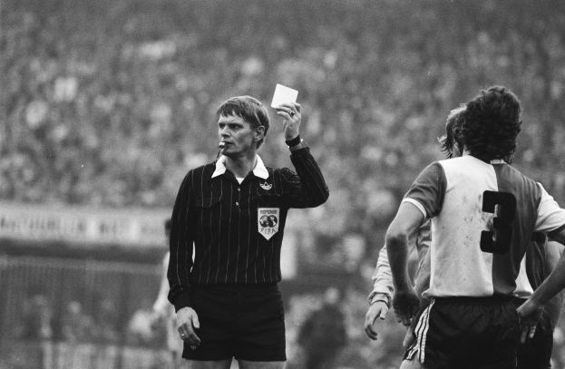 Schwarzes und weißes Bild eines Schiedsrichters, der eine rote Karte vor einer Menge von Menschen mit Fußballuniformen hält, mit einem Zaun im Hintergrund.