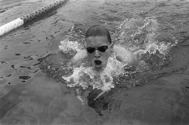 Ein Mann schwimmt in einem Pool mit offenem Mund und trägt eine Schwimmbrille, umgeben von Bahnmarkierungen während eines Rennens, dargestellt in Schwarz-Weiß.