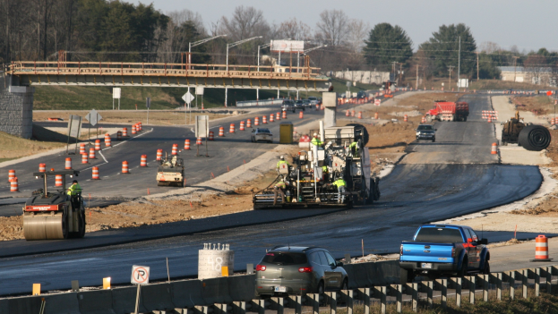 Baustelle mit Fahrzeugen auf der Straße, Verkehrsleitkegeln, Hinweistafeln, Masten, einer Brücke, Bäumen und einem klaren blauen Himmel.