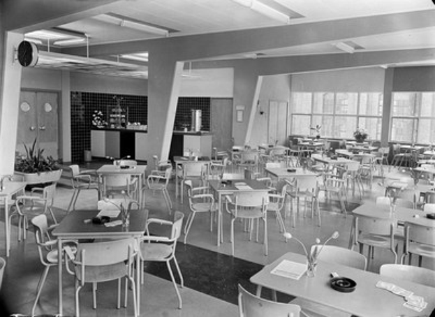 Schwarzes und weißes Foto einer Cafeteria mit Tischen und Stühlen, Tellern, Besteck, Gläsern und Blumenvasen auf den Tischen sowie Türen, Fenstern und Deckenlampen im Hintergrund.