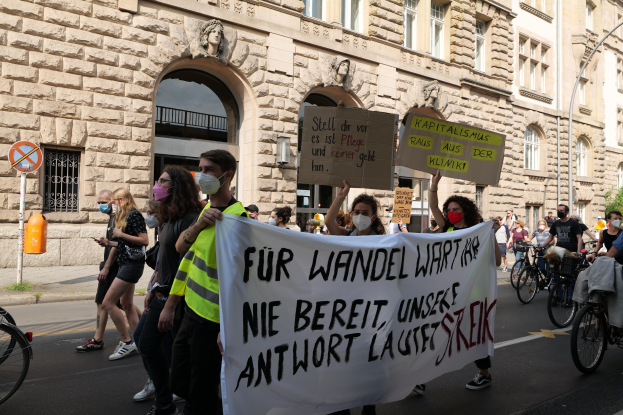 Eine Gruppe von Menschen marschiert auf einer Stadtstraße, einige halten Schilder und andere fahren Fahrräder, im Hintergrund ein Gebäude mit Fenstern, Bögen, Säulen und Skulpturen.
